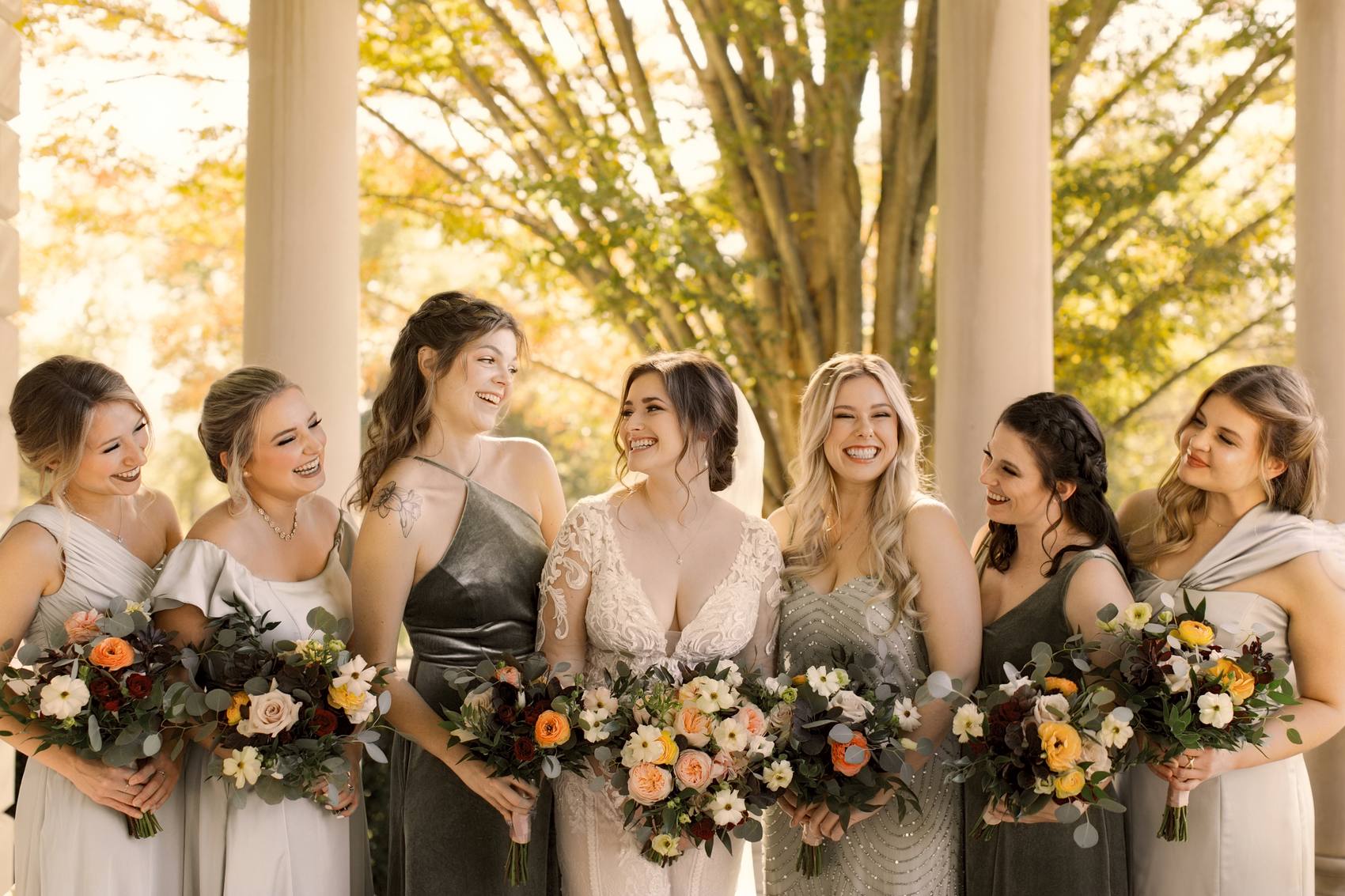 The bride laughing with six bridesmaids in sage, grey and velvet green gowns, each holding an autumn bouquet, between stone columns