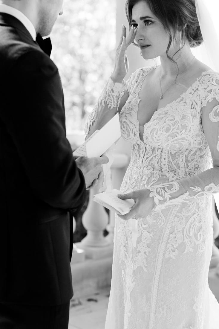 Black and white close photo of the bride lifting her hand to her face while the groom reads, her lace sleeves and veil framing her