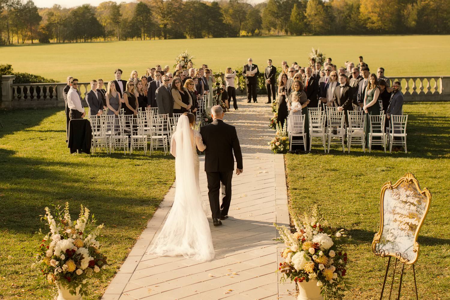 View from behind of the bride and a man in a black suit walking up a stone aisle toward guests, flower urns beside the path, a wide lawn and autumn trees beyond