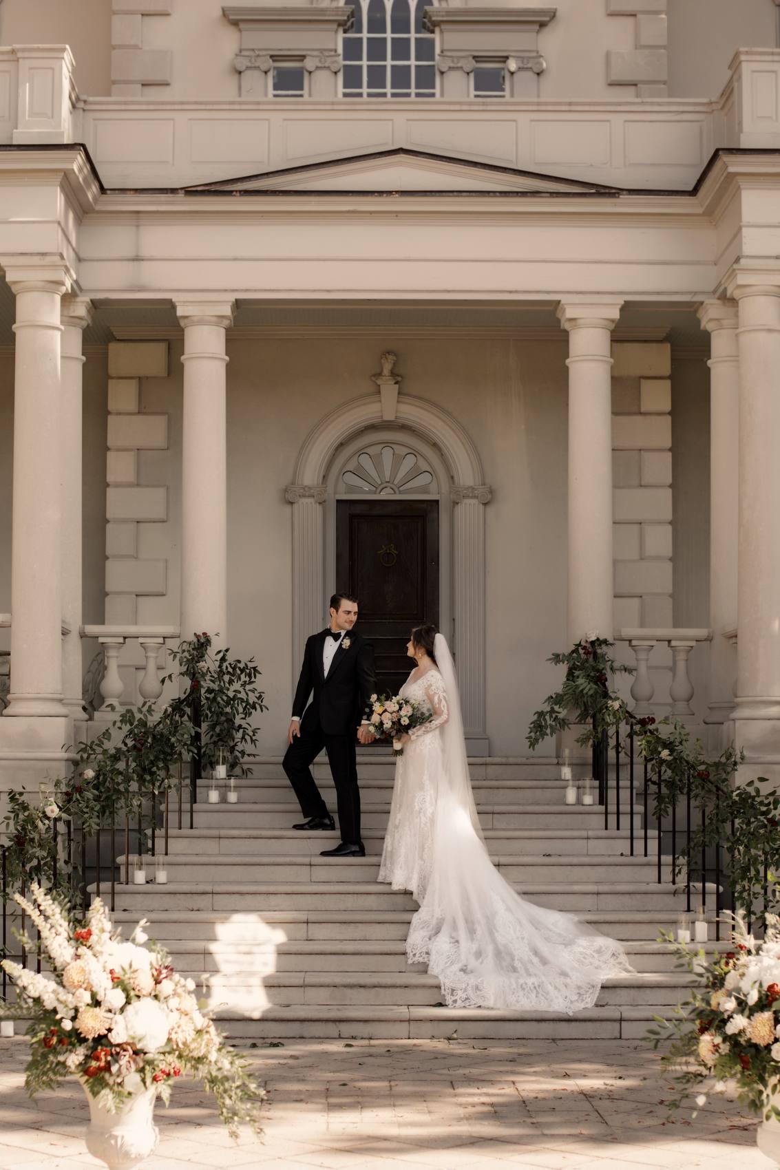 Groom looking back at the bride as they climb the front steps of a columned mansion, her long lace train and veil spread across the stone, urns of white and rust flowers in the foreground