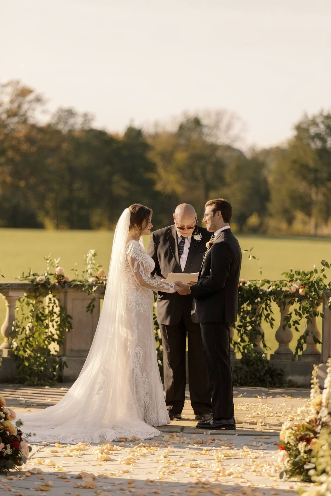 Bride and groom holding hands as they exchange vows before an officiant on a stone terrace, garlanded balustrade, a wide lawn and rose petals around them