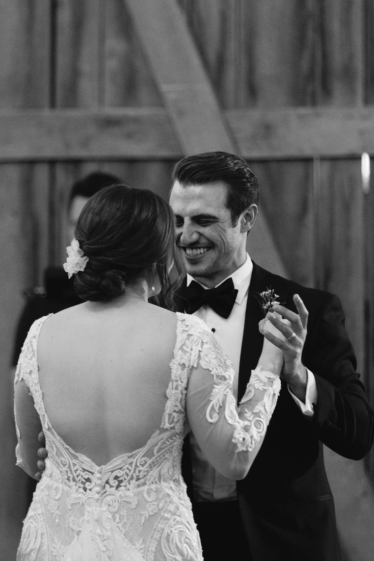 Black and white photo of the groom laughing as he dances with the bride, her open lace back and hair flower toward the camera, a wooden barn wall behind them