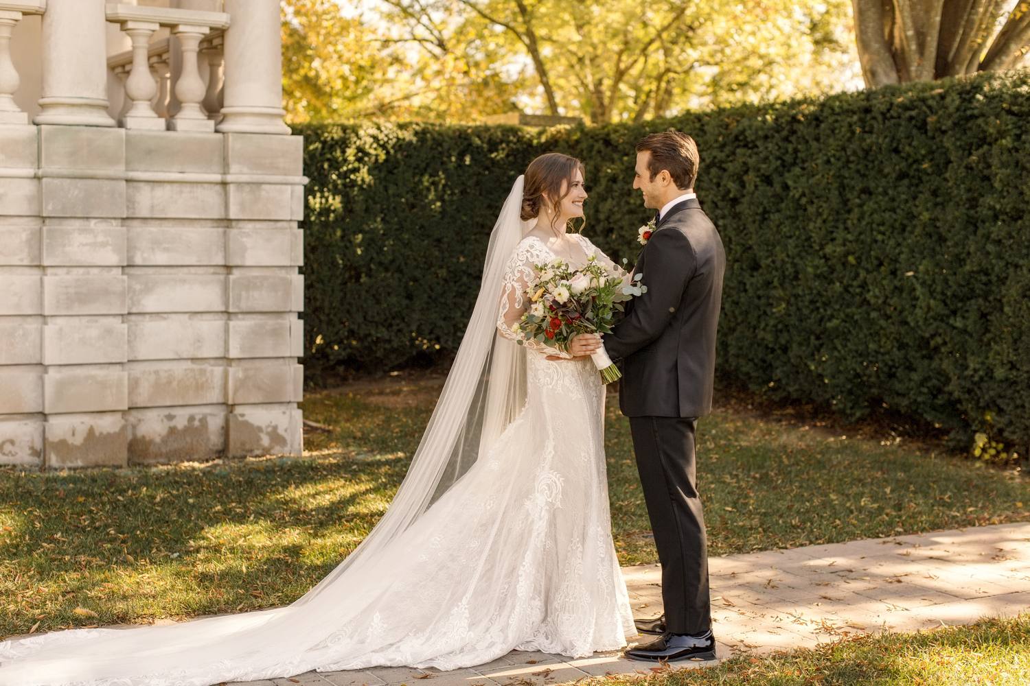 Bride with a very long veil facing the groom on a brick path beside a clipped hedge and a stone pillar, autumn light on the grass