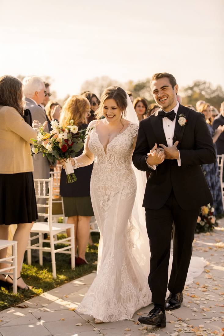 Bride and groom arm in arm and laughing as they walk up the aisle past clapping guests