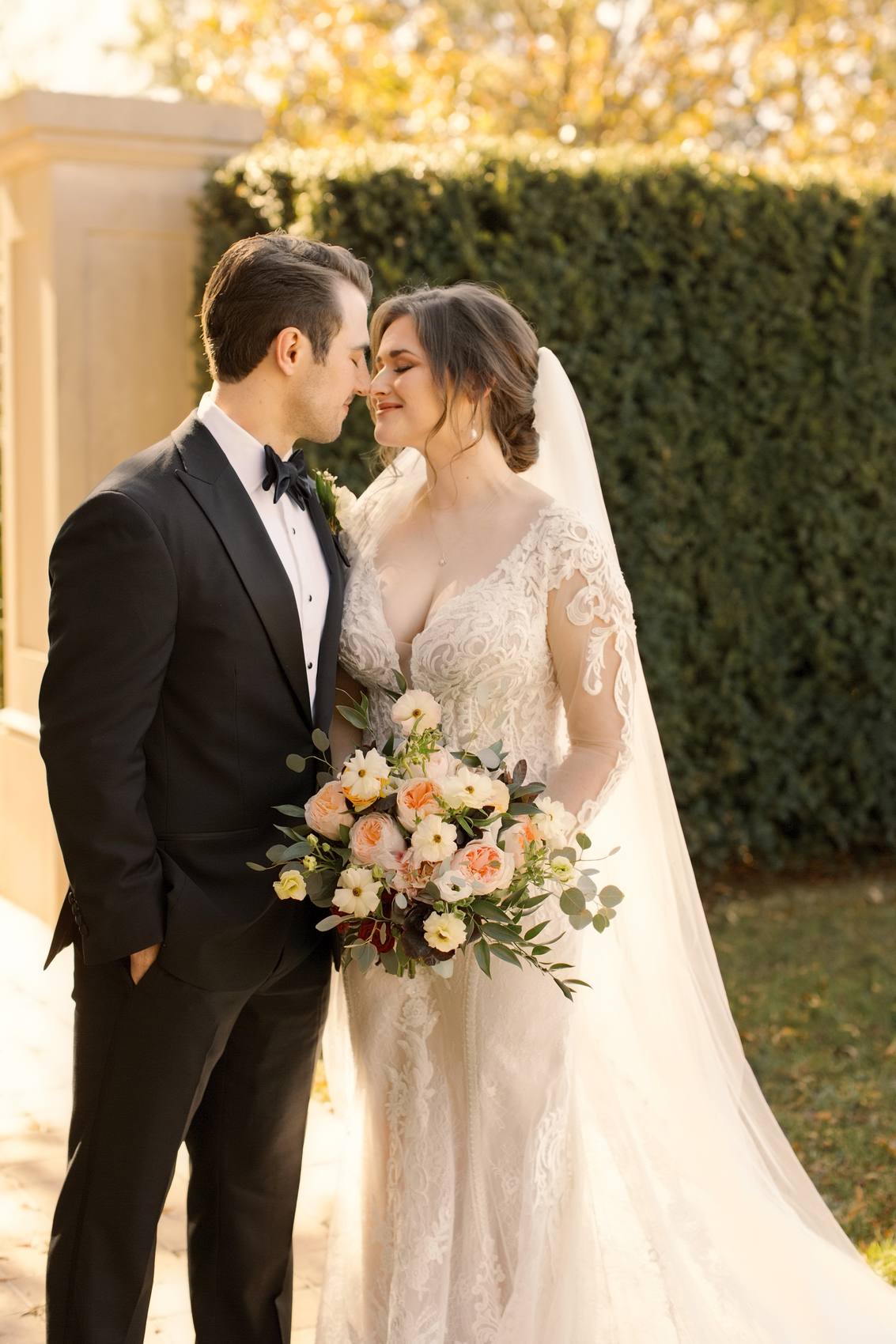 Groom in a black tuxedo and bride in a long veil and lace gown with their noses almost touching, her peach and cream bouquet held low, a clipped hedge and stone pillar behind them