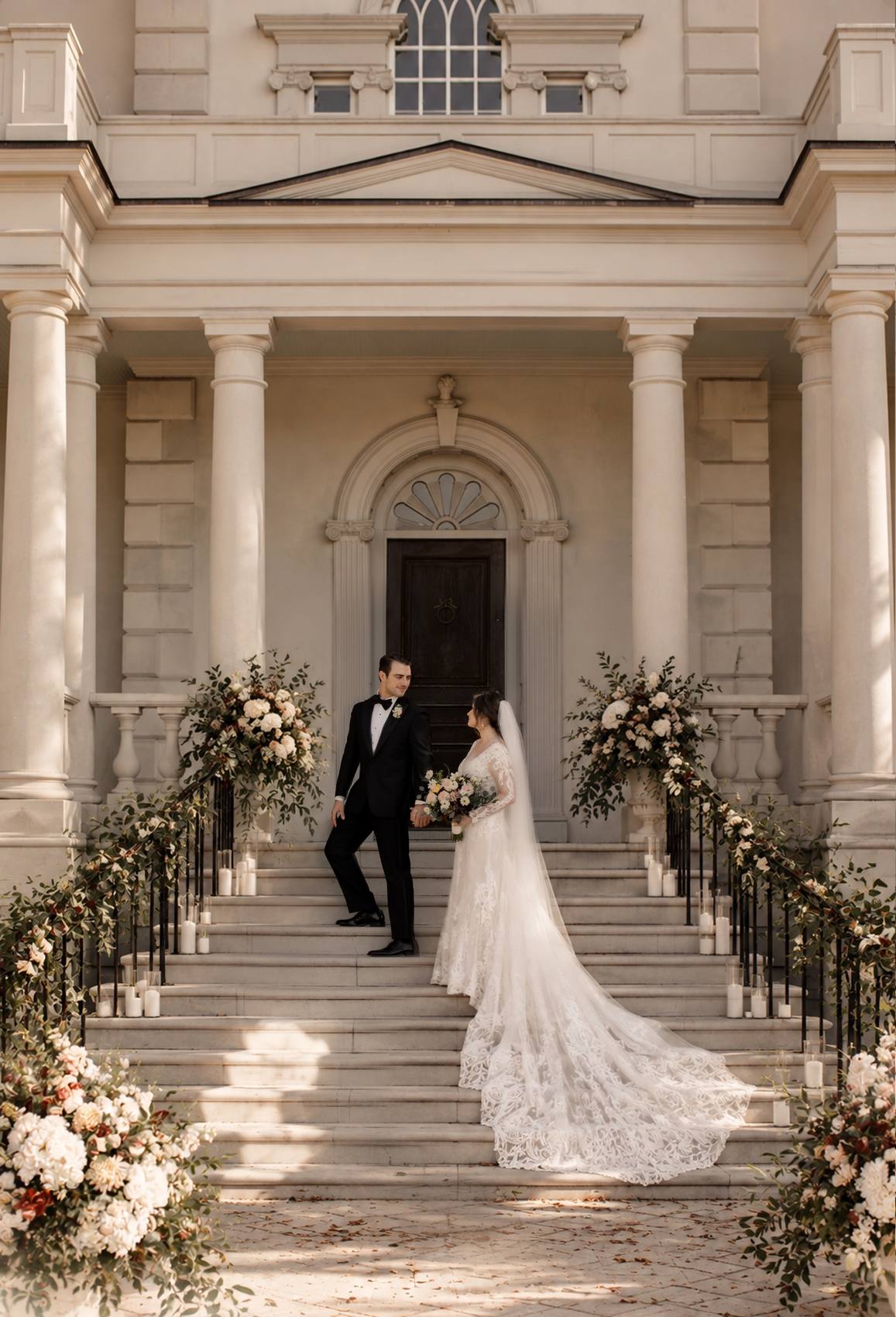 Groom looking back at the bride as they climb the front steps of a columned mansion, her long lace train spread across the stone, flower urns and candles along the garlanded railings