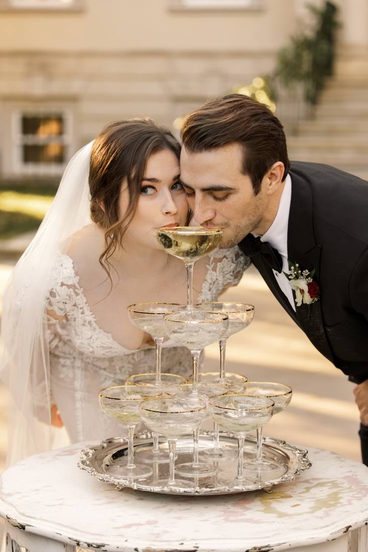 Bride and groom leaning in to drink from the top coupe of a gold-rimmed champagne tower on a distressed white table