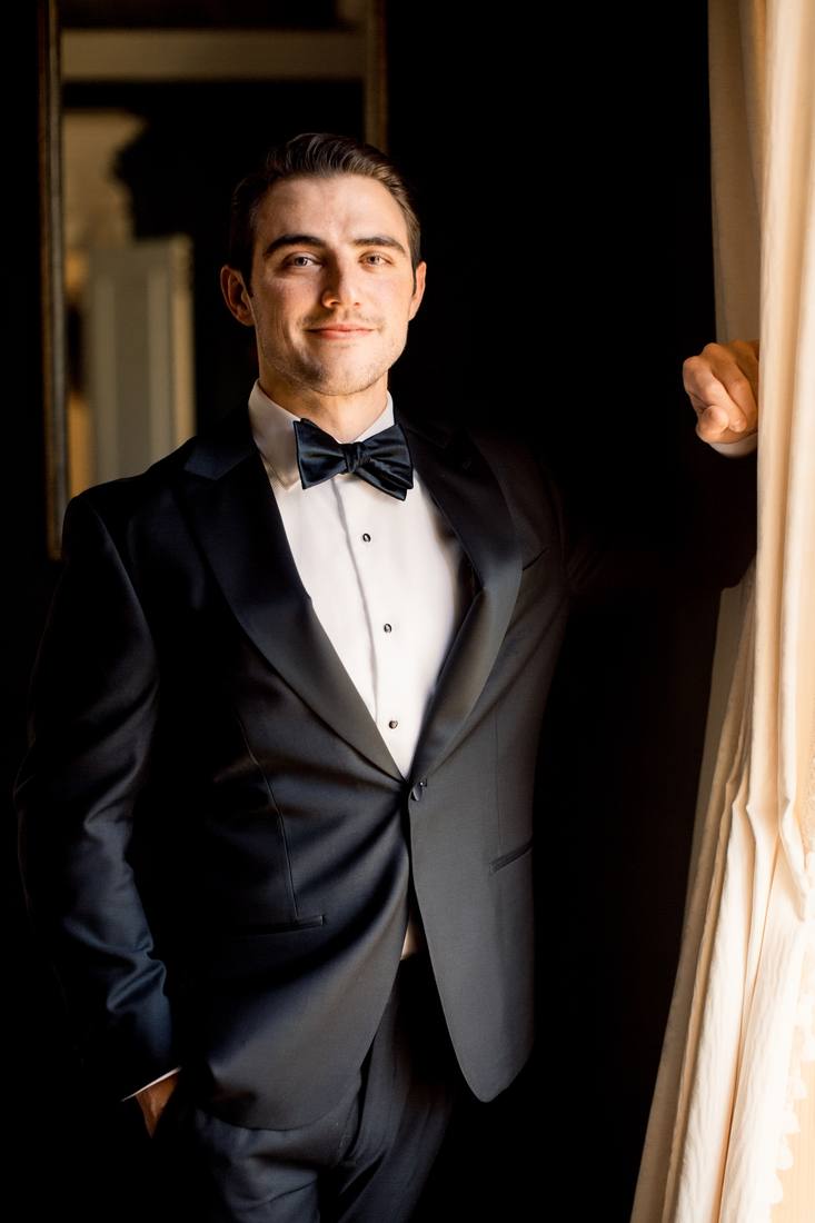 Groom in a black tuxedo and bow tie standing by a window curtain with one hand in his pocket