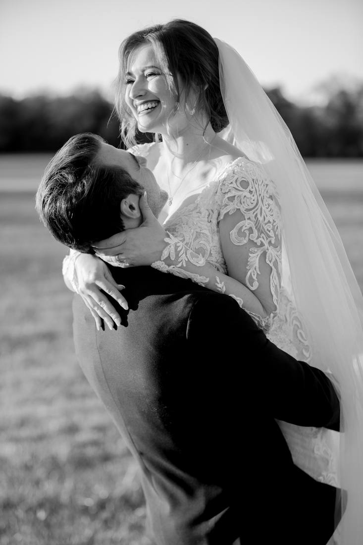 Black and white photo of the groom lifting the laughing bride, her lace sleeves and veil lit by low sun, an open field behind them