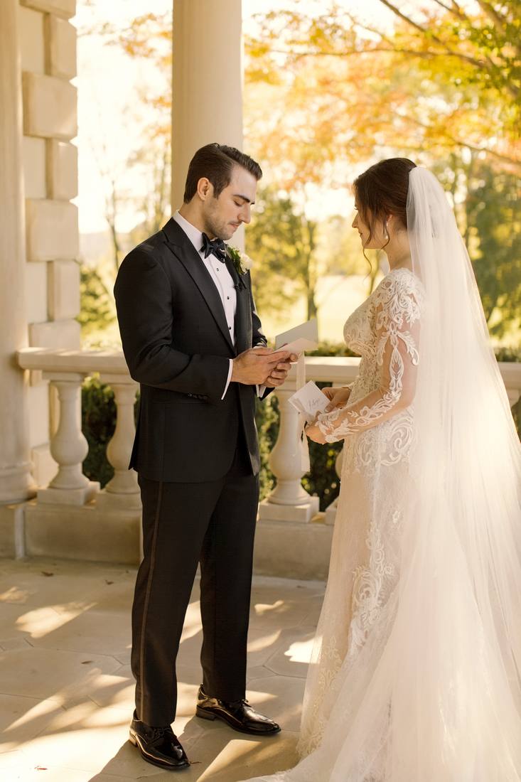 Groom reading his vows from a card to the bride between stone columns, autumn trees glowing behind them