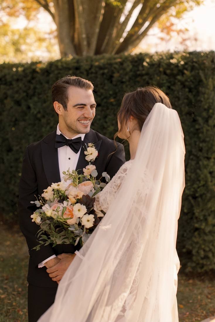 Groom smiling as he looks at the bride, her veil and peach bouquet in the foreground, a clipped hedge and autumn leaves behind them