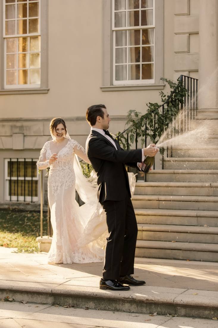 Groom spraying champagne from a bottle as the bride laughs behind him, her veil lifted by the wind, in front of mansion steps with a garland on the railing