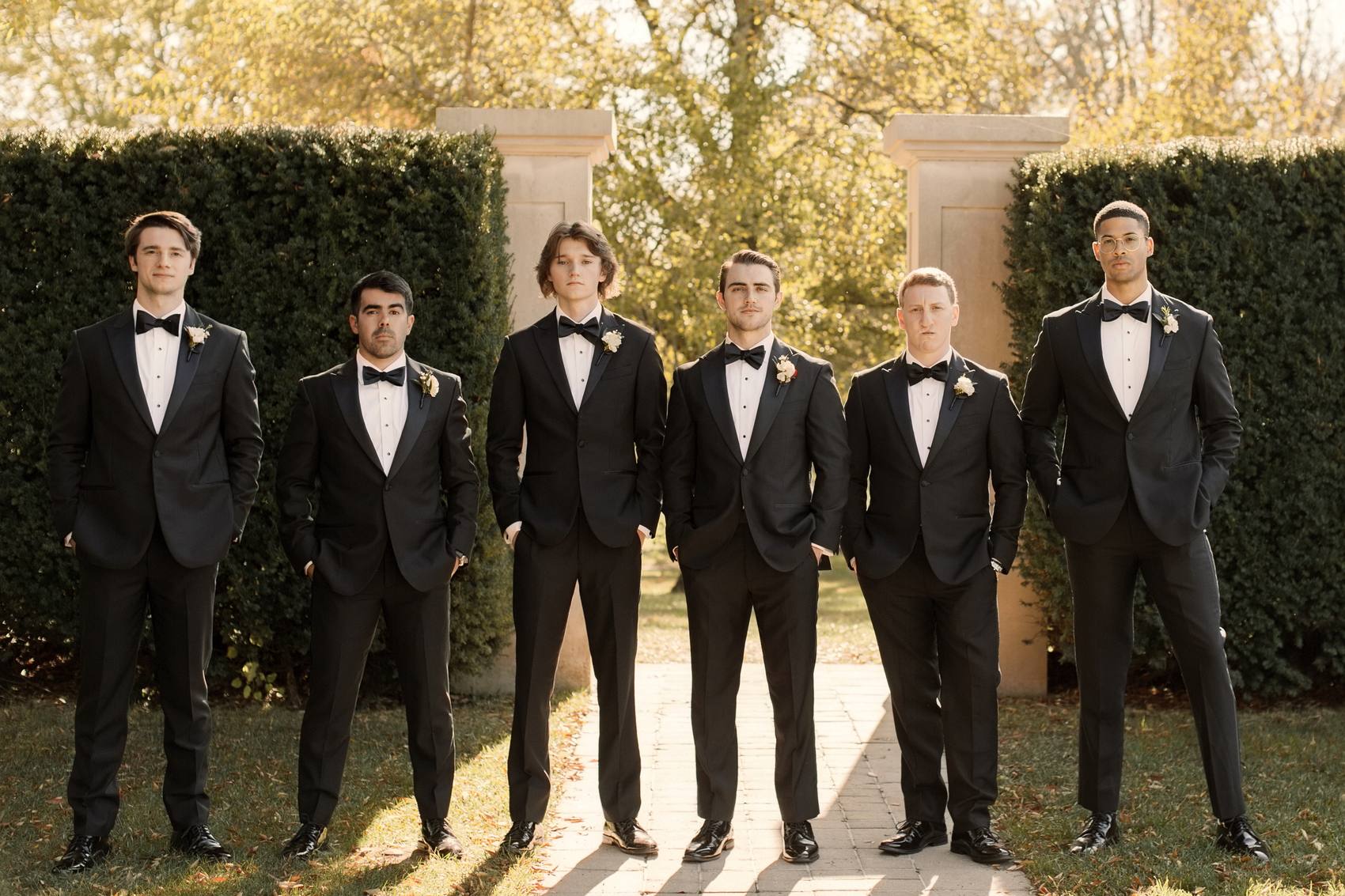 Six groomsmen in black tuxedos and bow ties standing in a line on a brick path between stone pillars and clipped hedges in warm autumn light