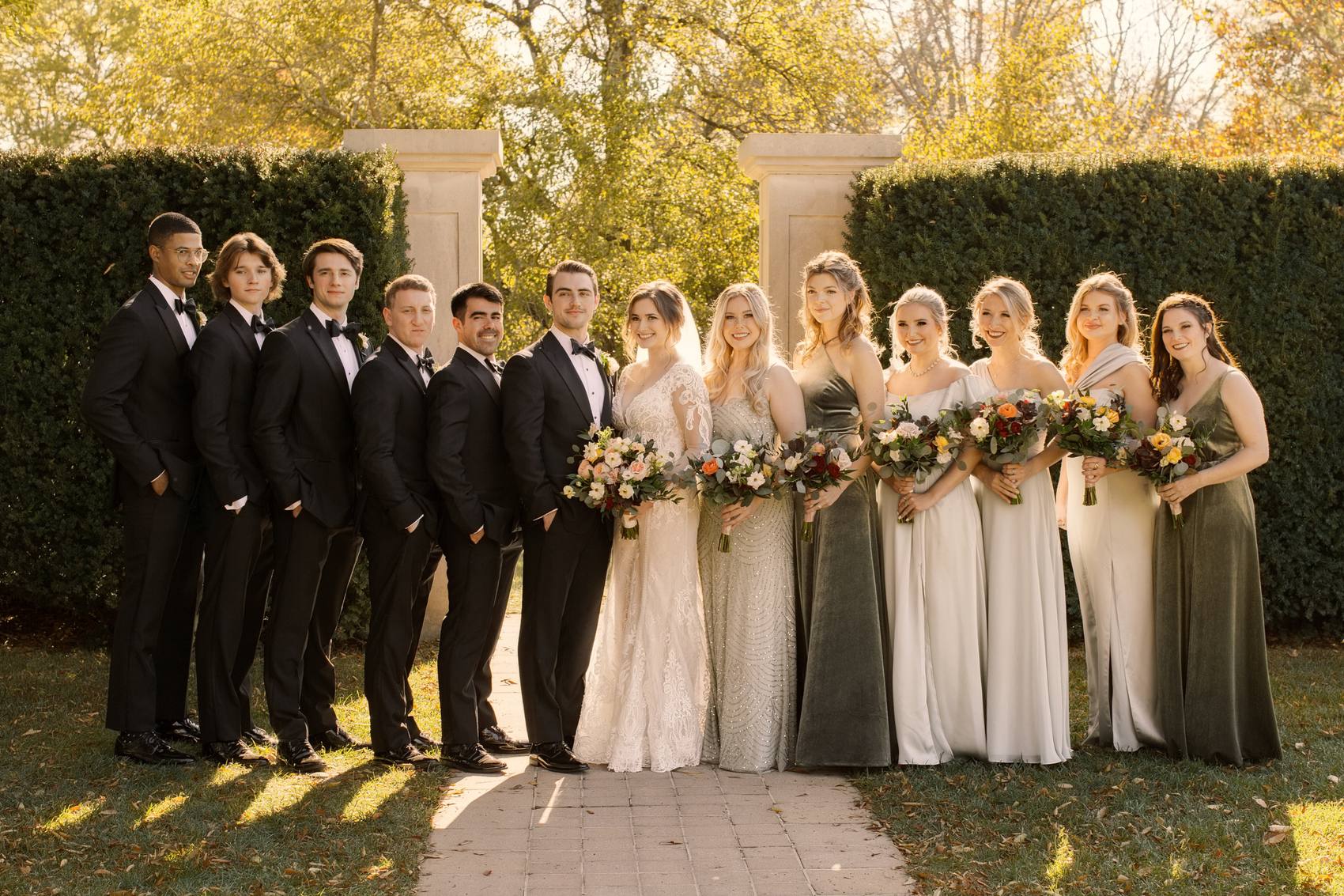 The bride and groom with their groomsmen and bridesmaids in sage and velvet green gowns, lined up on a brick path in front of stone pillars and hedges
