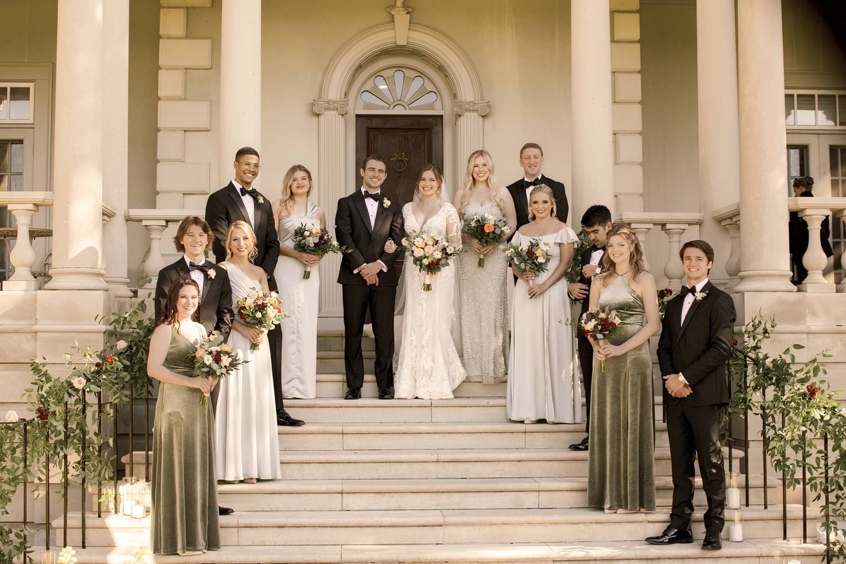 The couple with their bridesmaids and groomsmen posed on the wide front steps of a columned mansion, garlands and candles along the railings