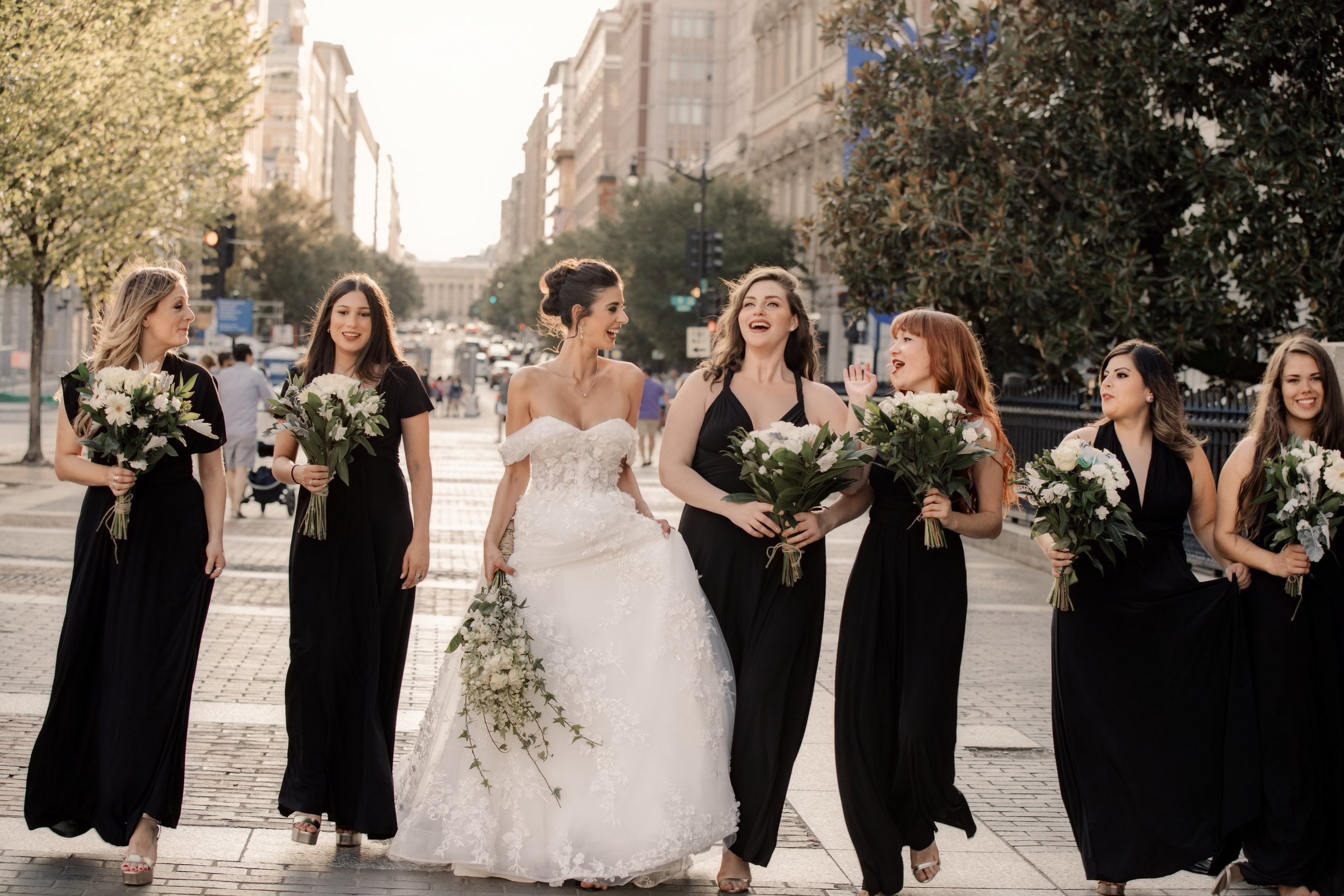 Bride and bridesmaids in black gowns walking toward the camera down a sunlit downtown Washington DC street, each holding white bouquets
