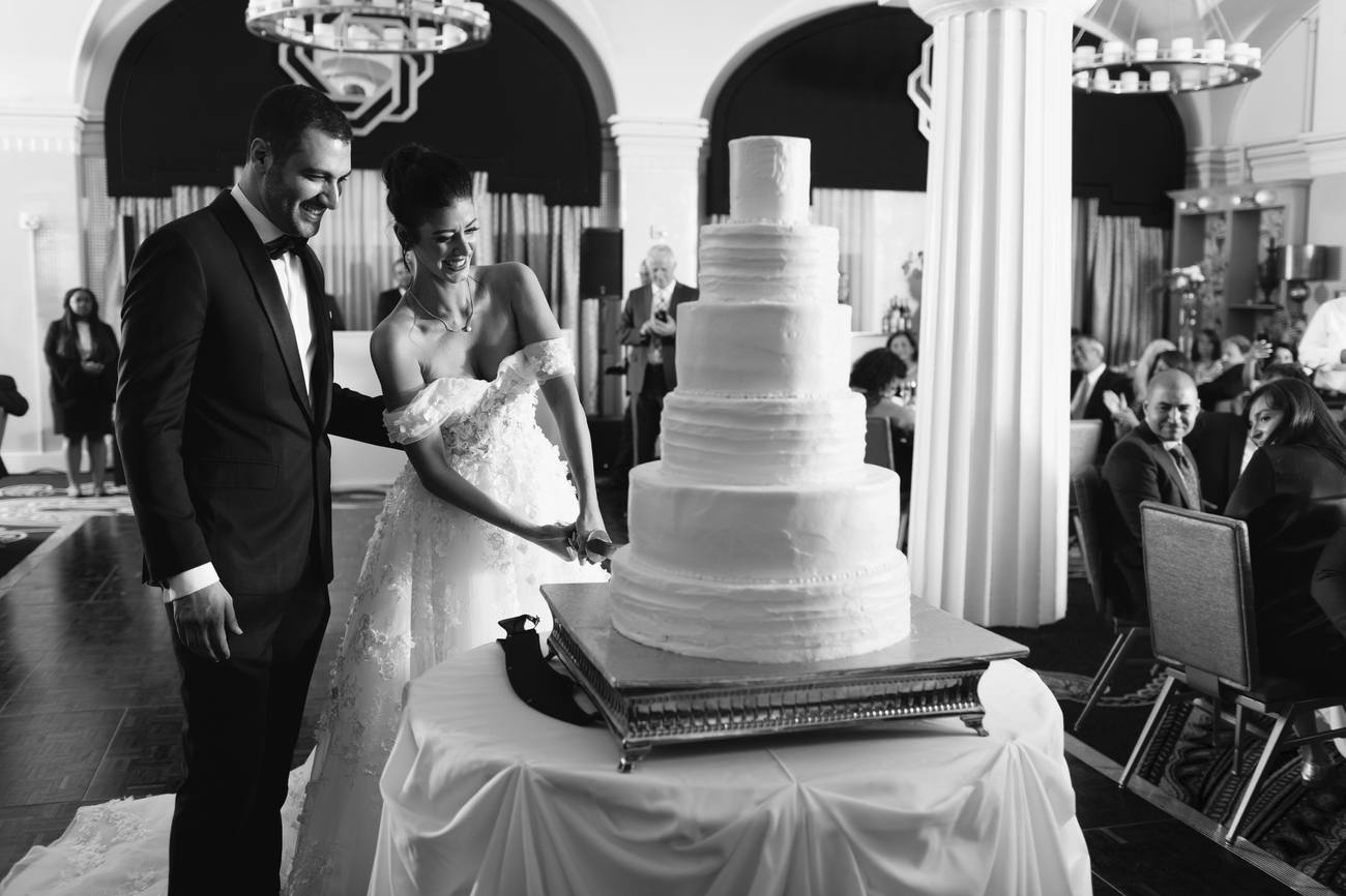 Black and white photo of the bride and groom cutting a tall white tiered wedding cake as guests watch