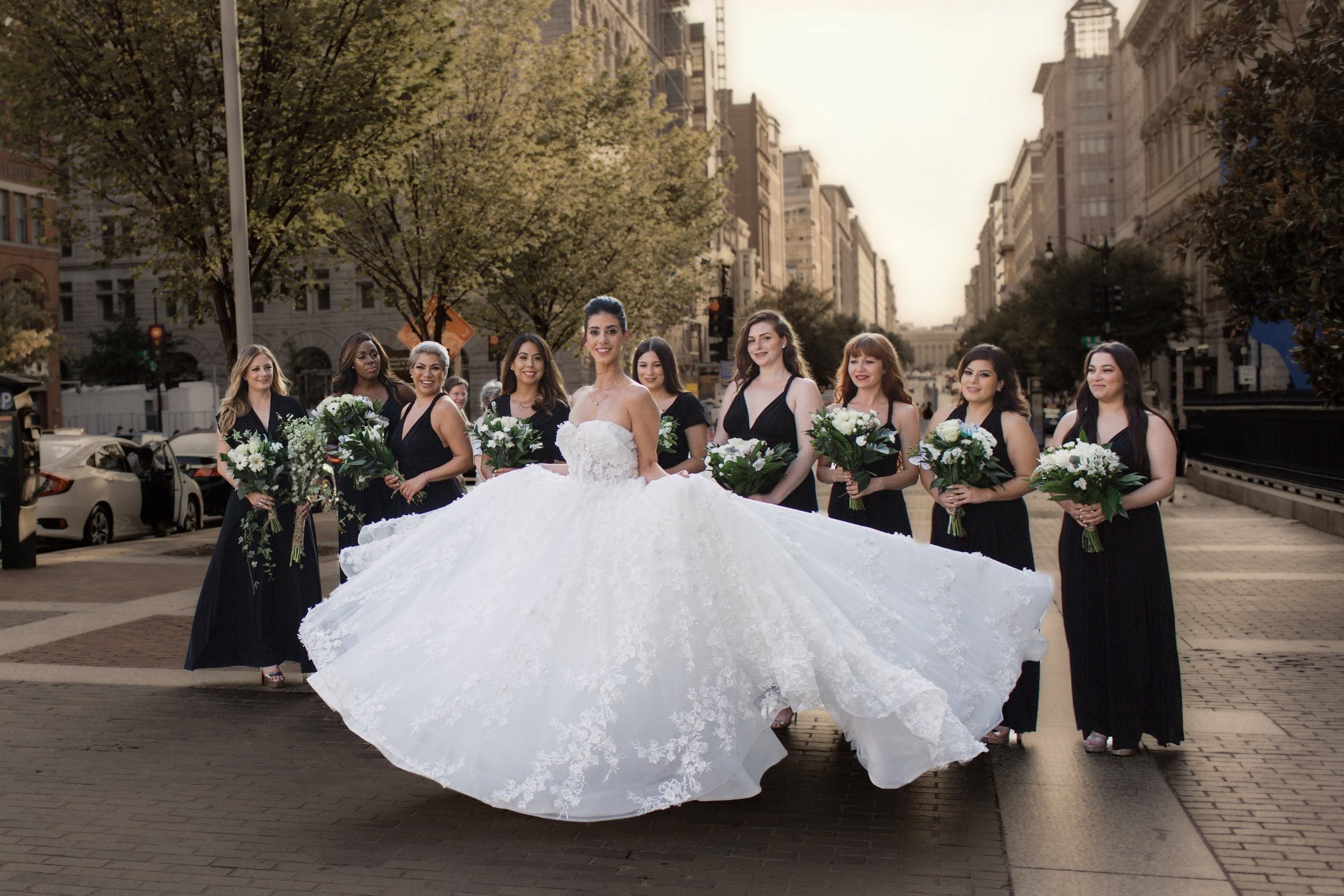 Bride in a voluminous lace gown twirling on a sunlit downtown Washington DC street, her skirt swept out as nine bridesmaids in black dresses with white bouquets stand behind her.