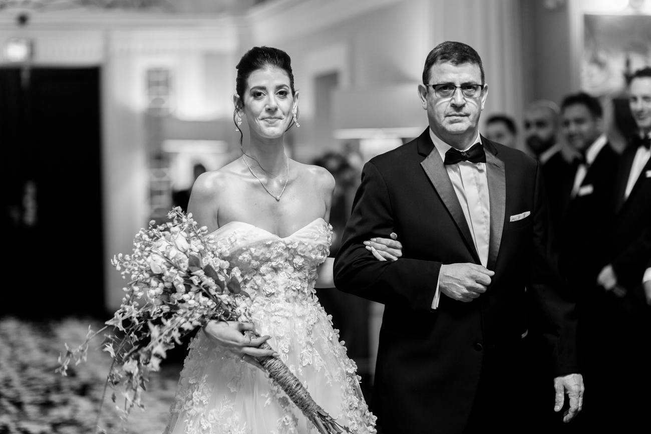 Black and white photo of a bride with a trailing bouquet walking arm in arm with a man in a tuxedo as guests look on