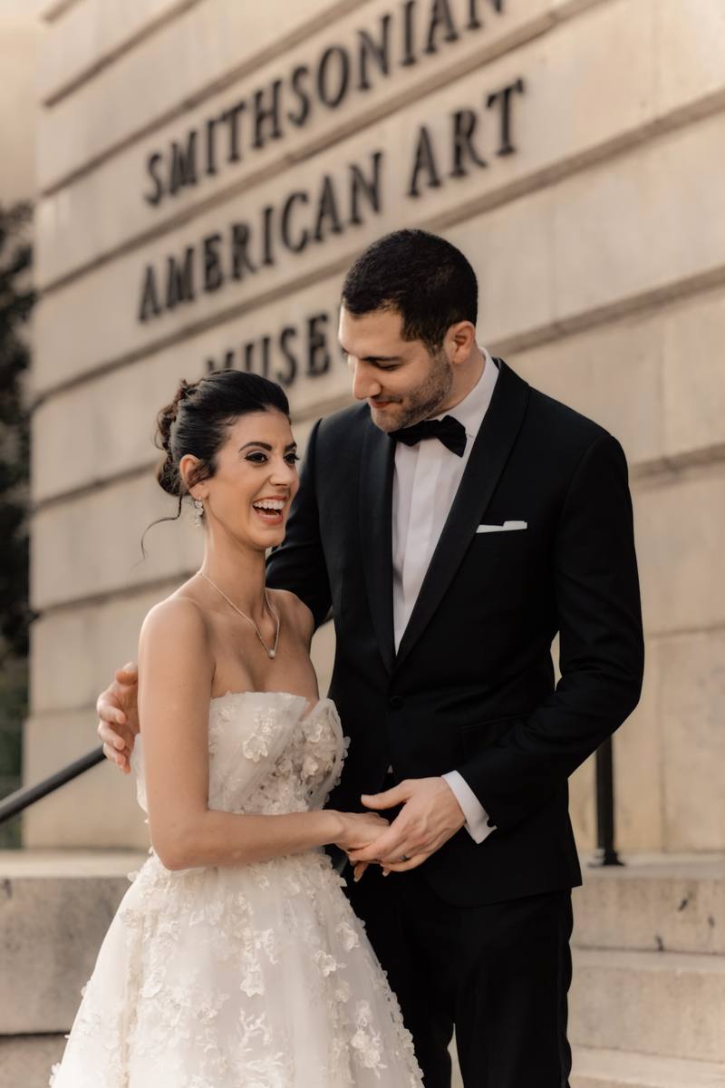 Bride laughing as the groom looks down at her in front of the Smithsonian American Art Museum sign on stone steps in Washington DC