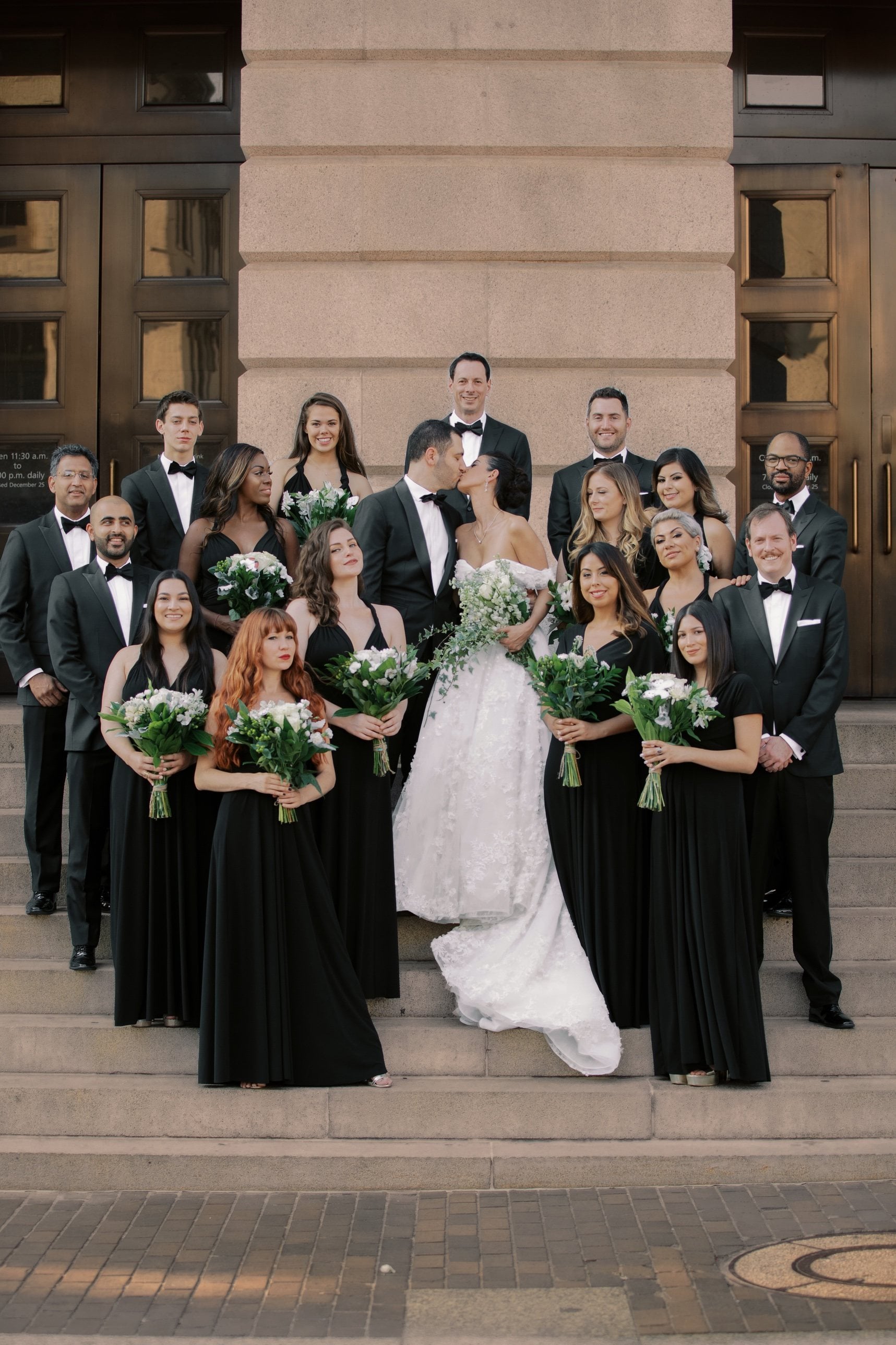 Wedding party of bridesmaids in black gowns with white bouquets and groomsmen in tuxedos on stone steps, the couple kissing at the center