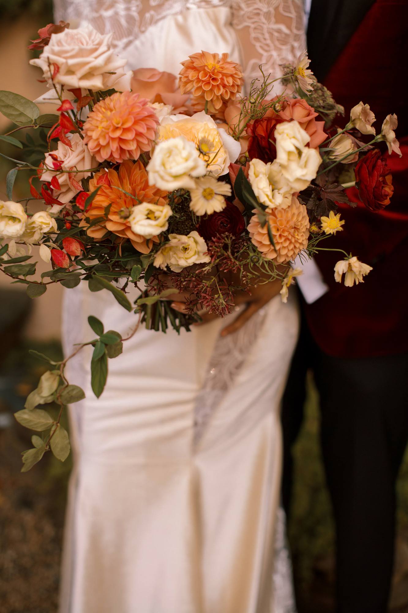 Close view of an autumn bouquet of orange dahlias, blush roses, cream lisianthus and burgundy ranunculus held against a lace and satin gown