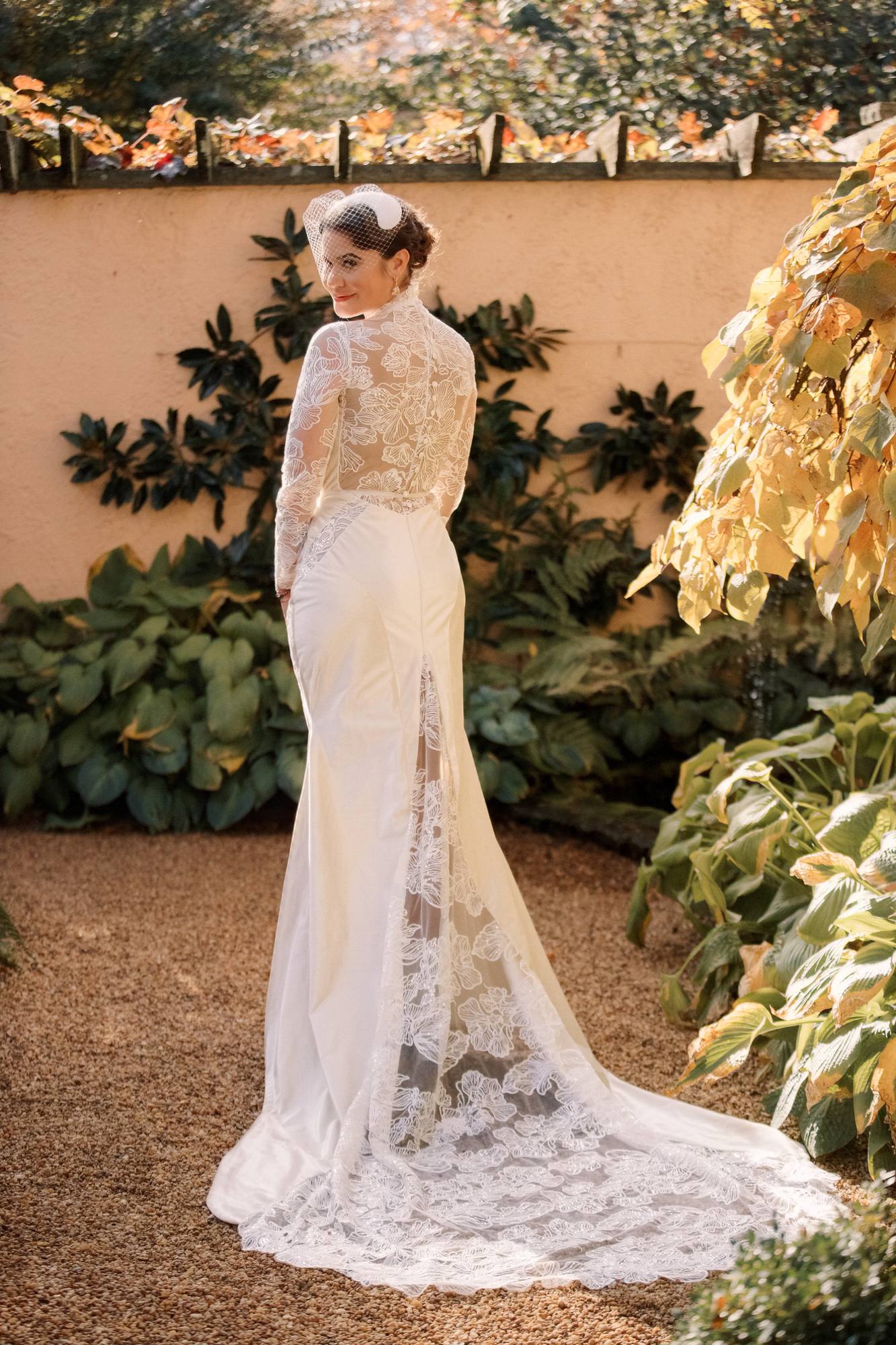 Bride seen from behind in a sheer lace-back gown with a long train, looking over her shoulder in a courtyard with yellow autumn leaves