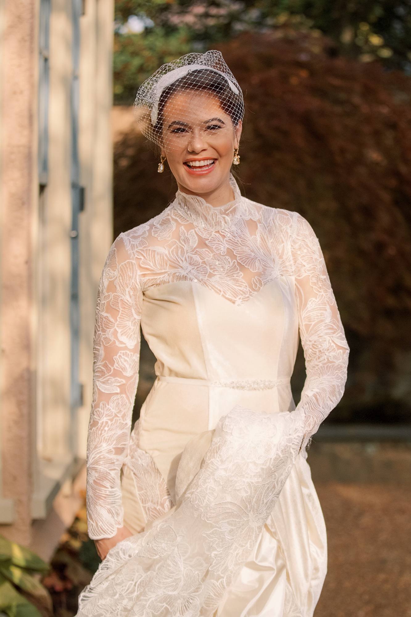 Bride laughing in a long-sleeved lace gown and a birdcage veil, holding her train in an autumn garden