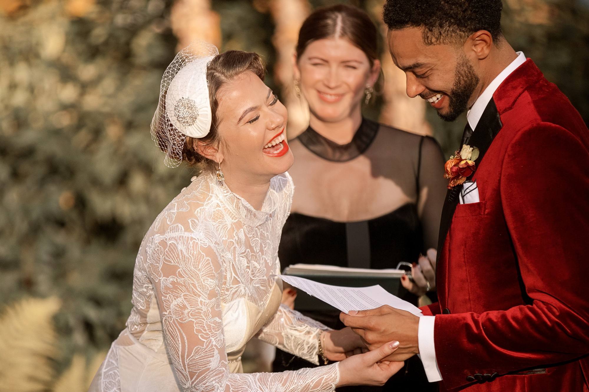 Bride laughing out loud as the groom reads his vows from a sheet of paper, the officiant smiling between them