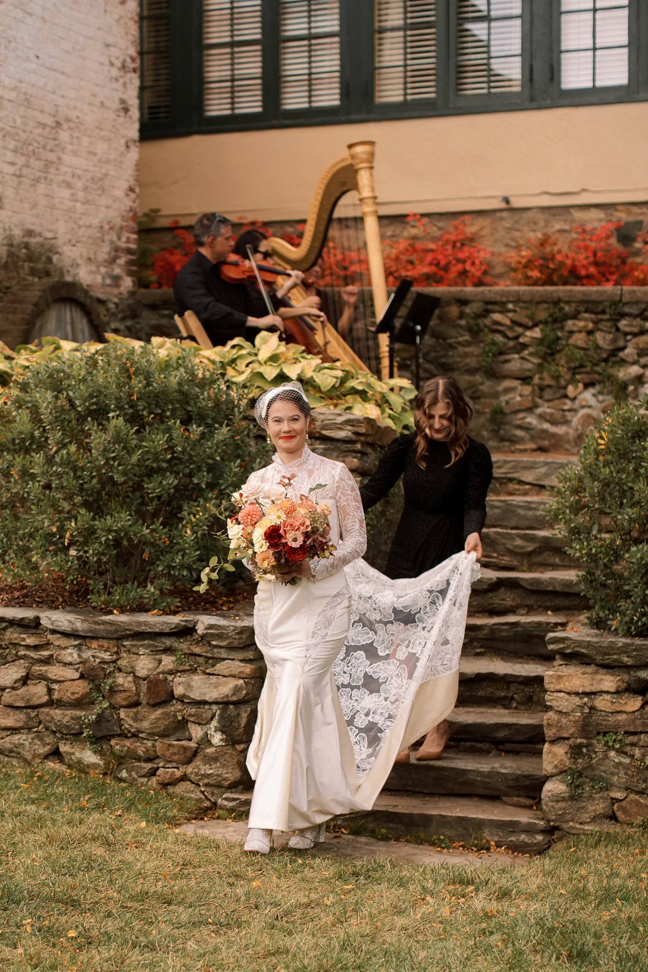 Bride in a lace gown carrying an autumn bouquet as she walks down stone steps while a woman in black lifts her lace train, a harp and string players behind