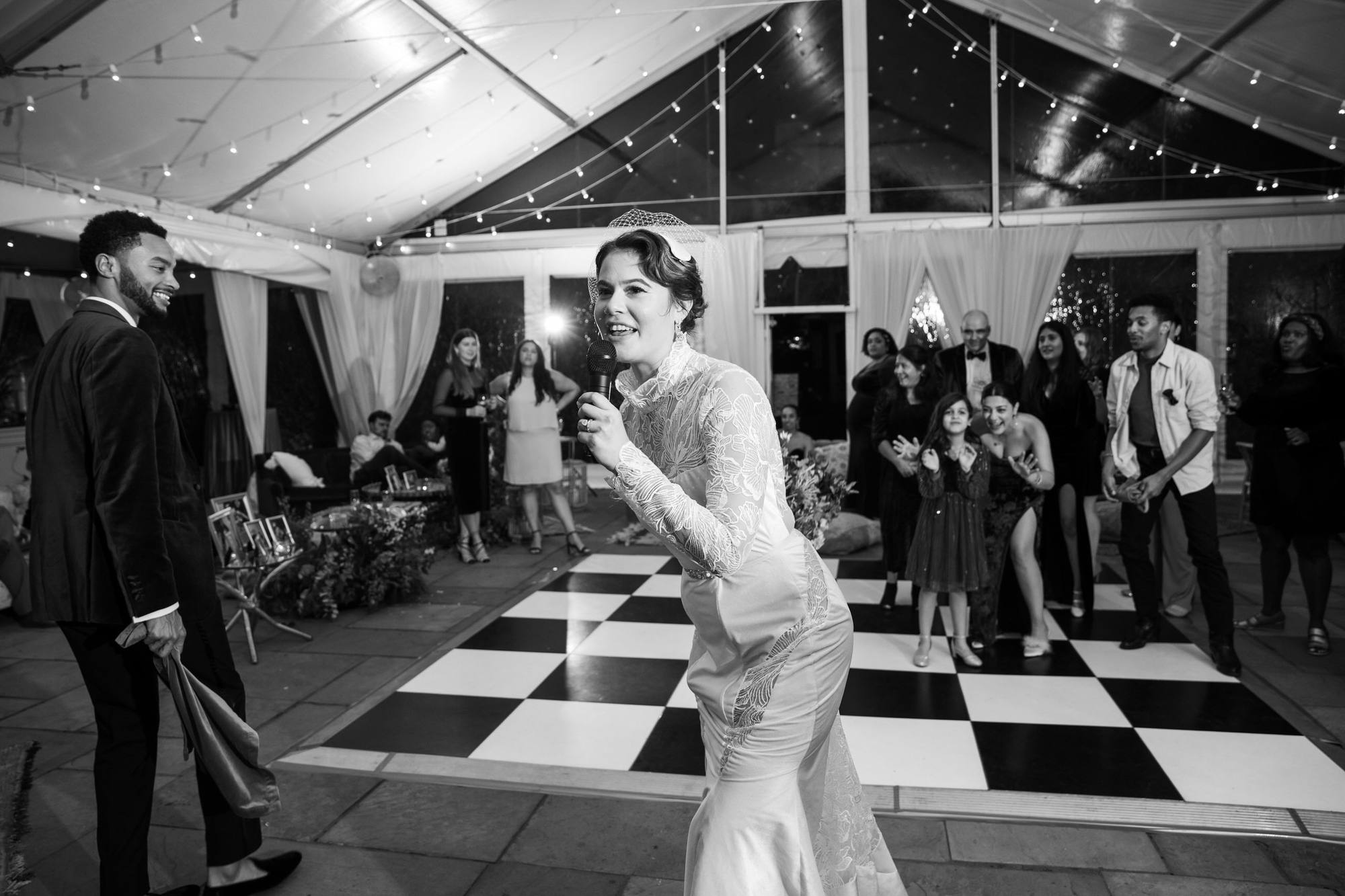 Black and white image of the bride speaking into a microphone on the dance floor as guests gather behind her and the groom looks on, under a clear tent hung with string lights