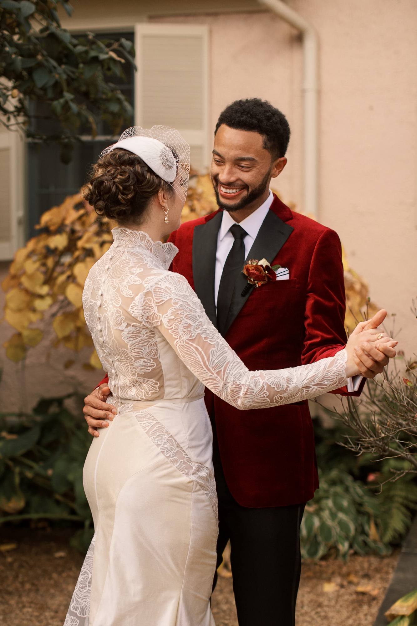 Bride in a lace gown and groom in a red velvet jacket holding hands in a dance pose, he laughing, in a courtyard with yellow autumn leaves