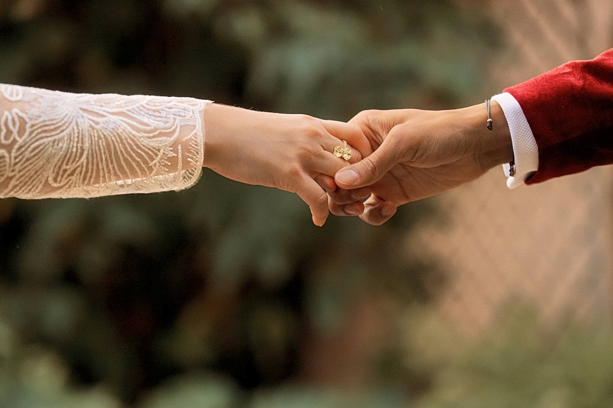 Close view of the couple holding hands, her lace sleeve and gold floral ring beside his red velvet cuff