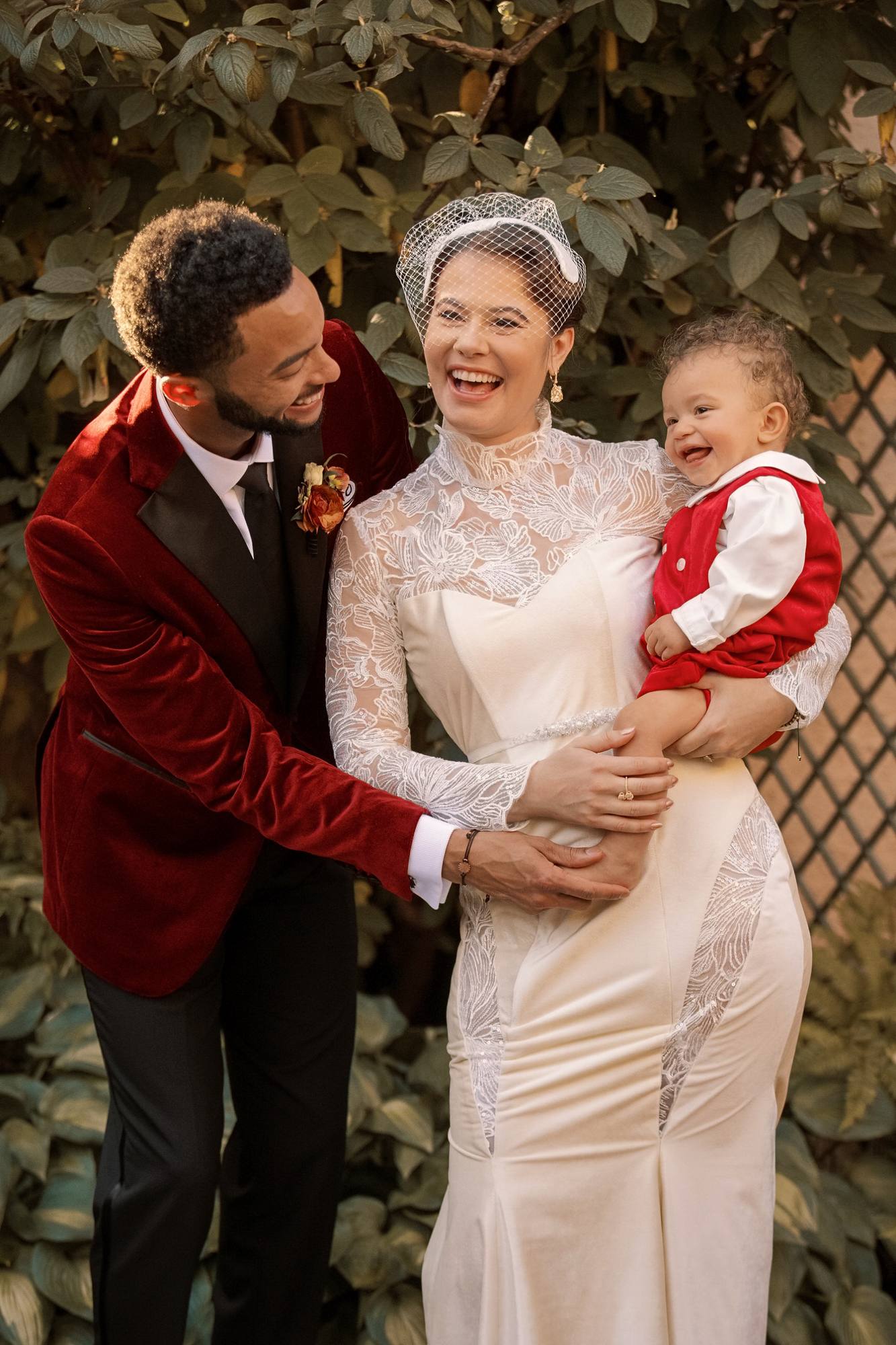 Groom, bride and their baby boy laughing together in a garden, the baby in a red velvet vest and overalls