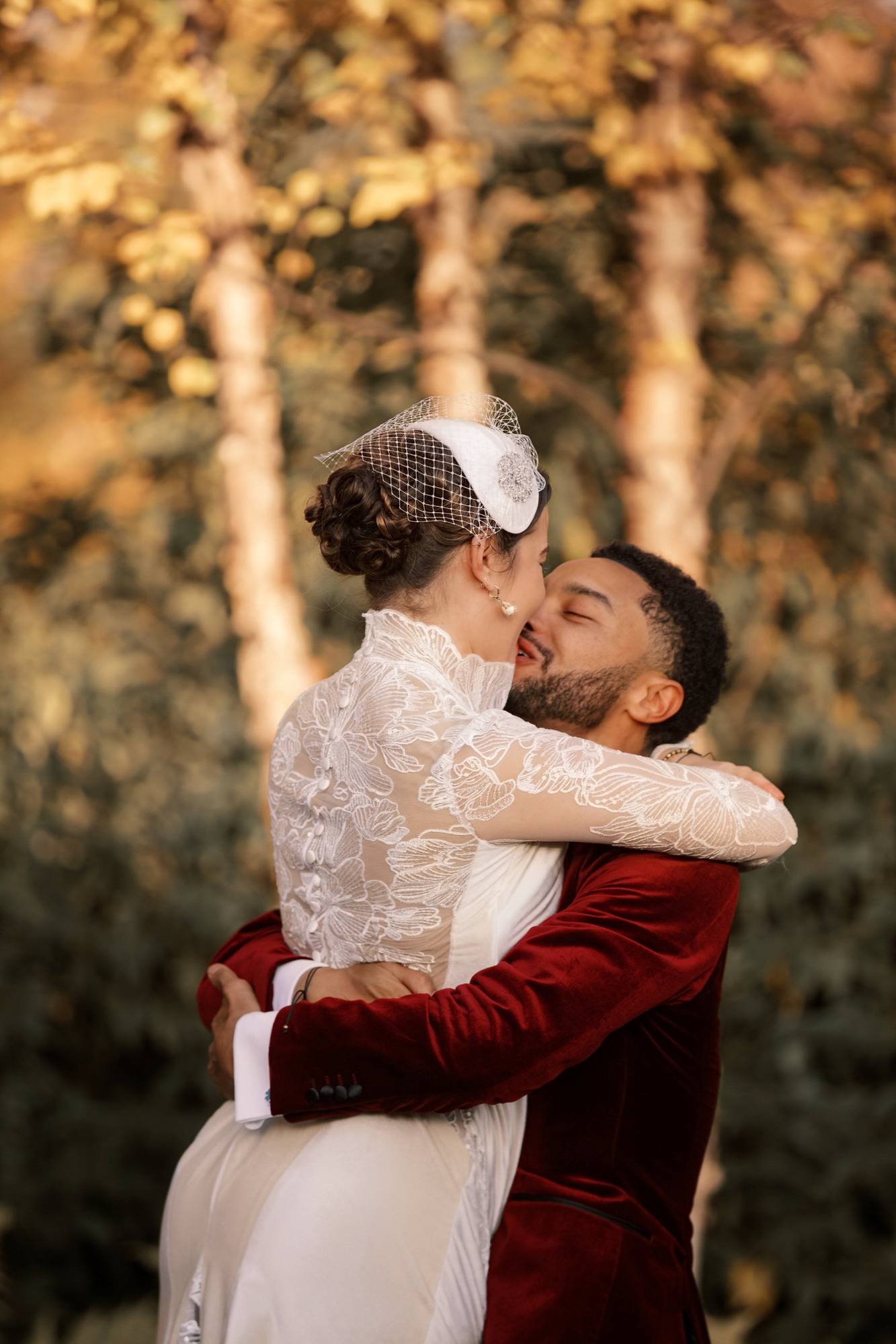 Couple embracing and kissing in front of birch trees with golden leaves, the bride's lace back and covered buttons toward the camera