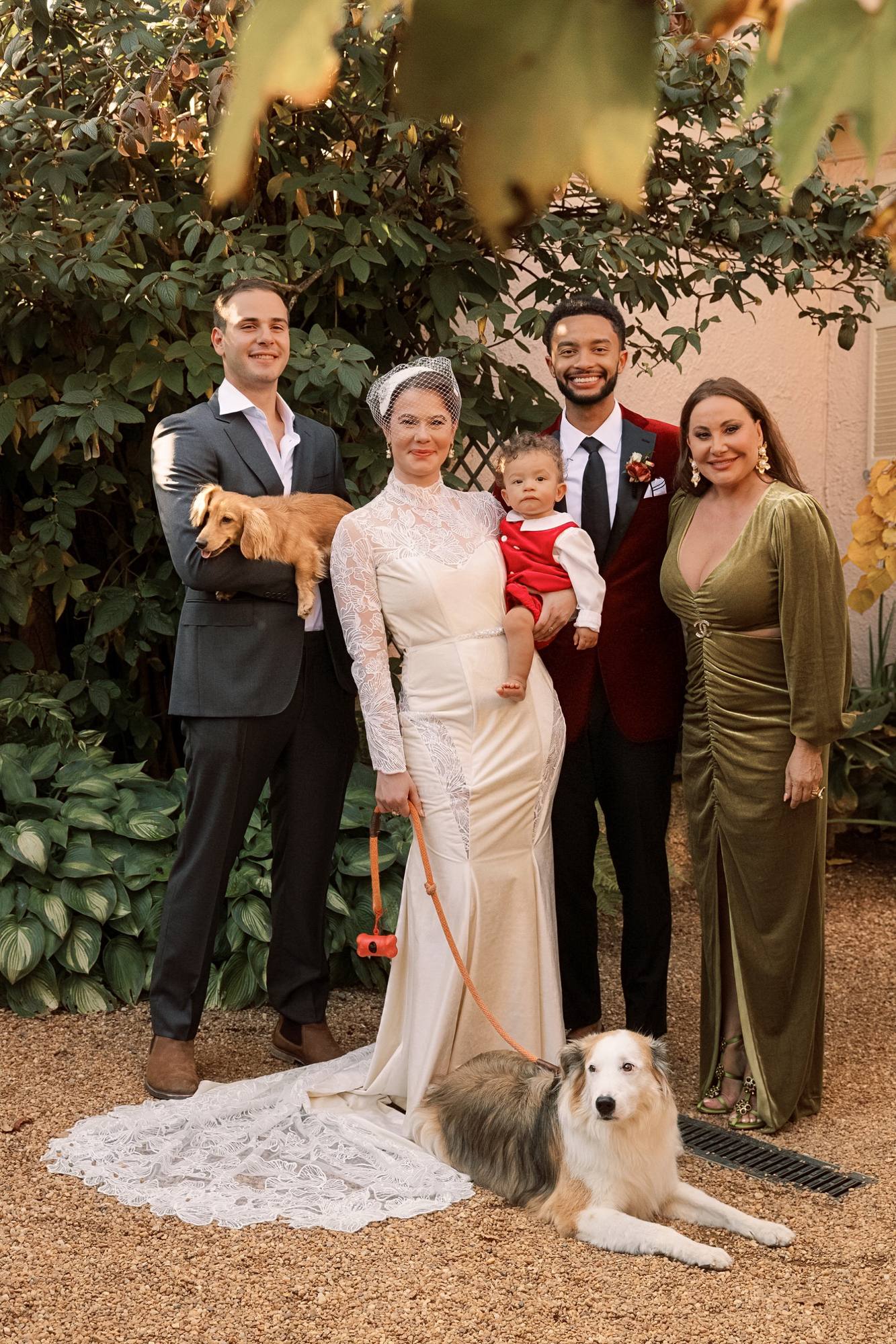 Family portrait on gravel with a man holding a dachshund, the couple with their baby, a woman in an olive velvet dress and a dog lying on the bride's lace train
