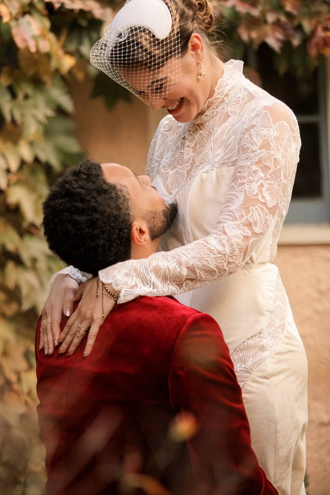 Bride laughing as the groom lifts her, in front of a stucco wall covered in autumn vines