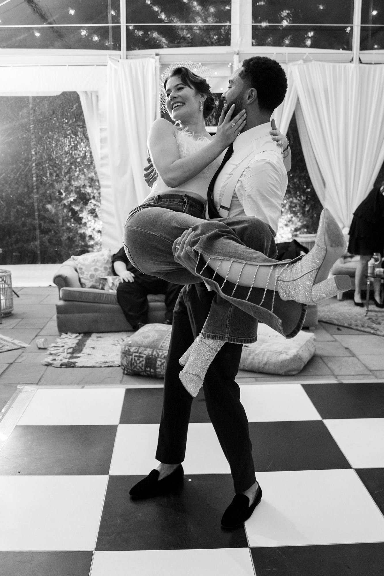 Black and white image of the groom carrying the bride, now in jeans, a feathered top and sparkly boots, on a checkered dance floor