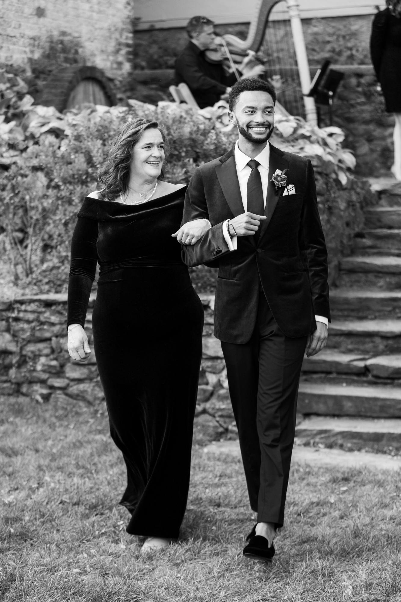 Black and white photo of the groom in a velvet tuxedo jacket walking arm in arm with a smiling woman in a black velvet gown, a harp and violinist behind them on a stone terrace