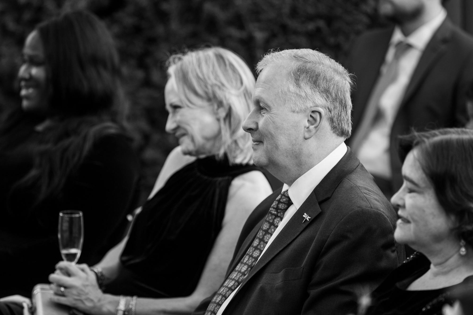 Black and white portrait of guests smiling during the ceremony, one holding a champagne flute and one wearing a dragonfly pin