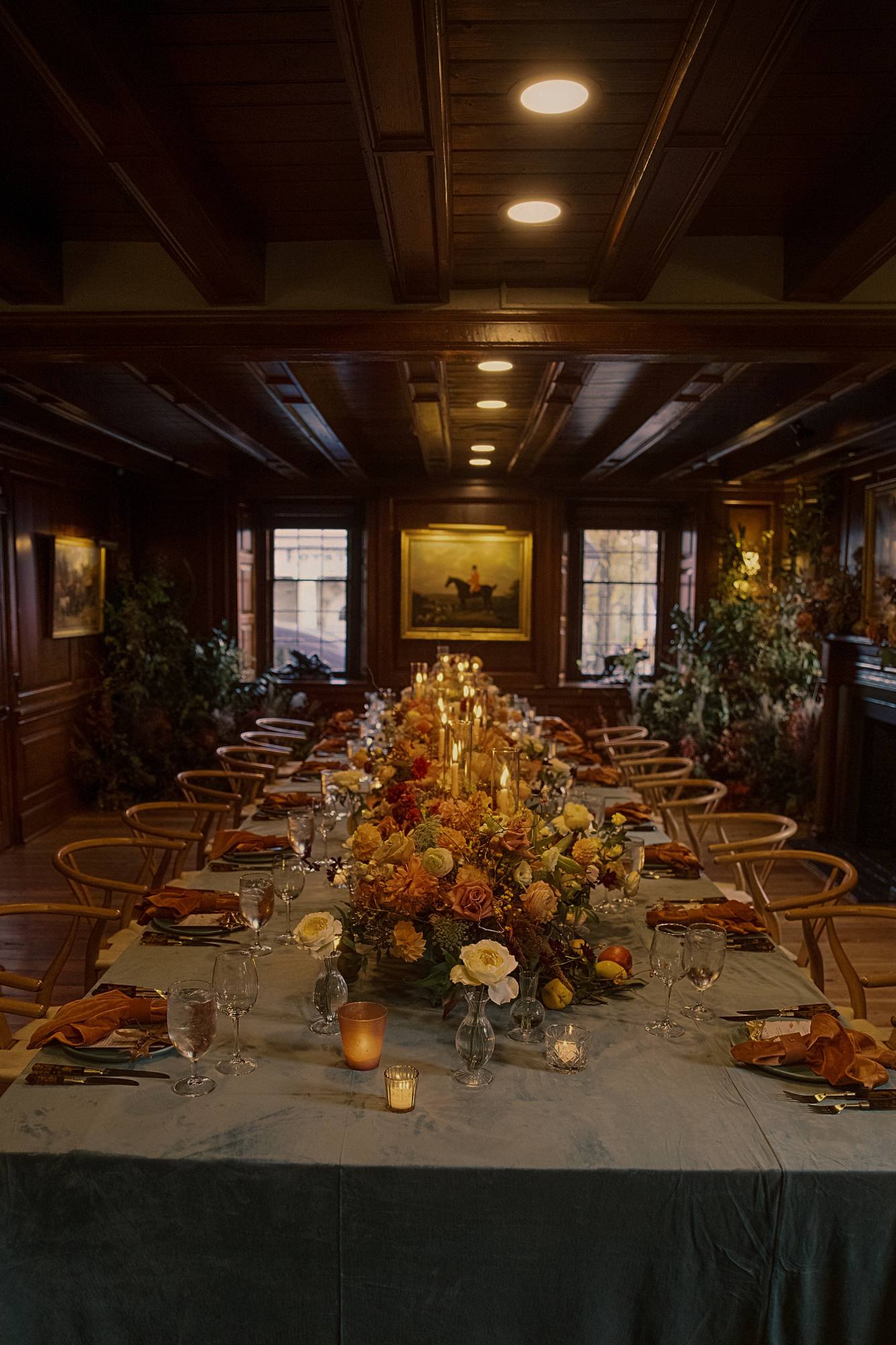 Long reception table in a wood-paneled room with a teal velvet cloth, wishbone chairs, candles and a low centerpiece of dahlias, roses and ranunculus, with a hunting portrait on the far wall