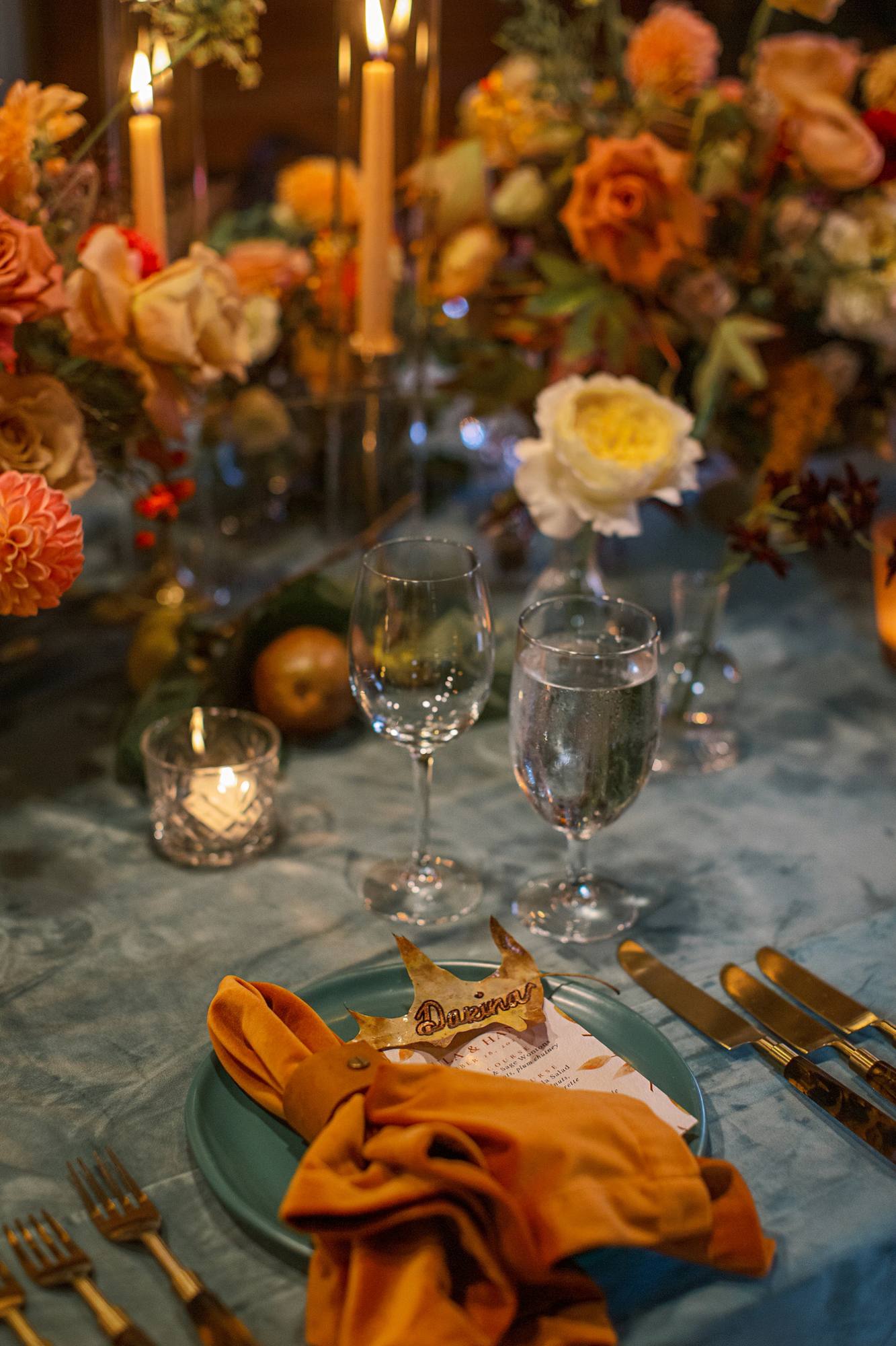 Reception place setting with a pressed autumn leaf place card, an orange velvet napkin, a teal plate and brass flatware on a teal velvet cloth