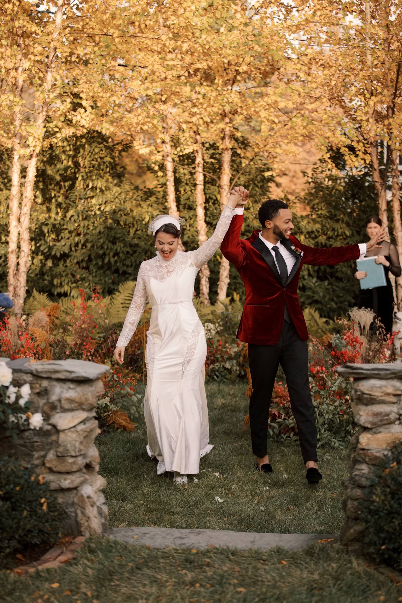 Couple walking up the aisle with joined hands raised between two stone pillars, birch trees in full gold behind them and the officiant at the right