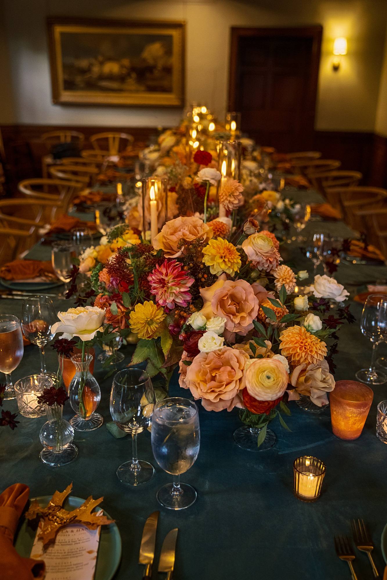 Close view of the table centerpiece of yellow and red dahlias, peach roses and ranunculus among taper candles and bud vases, with a menu on an autumn leaf at the lower left