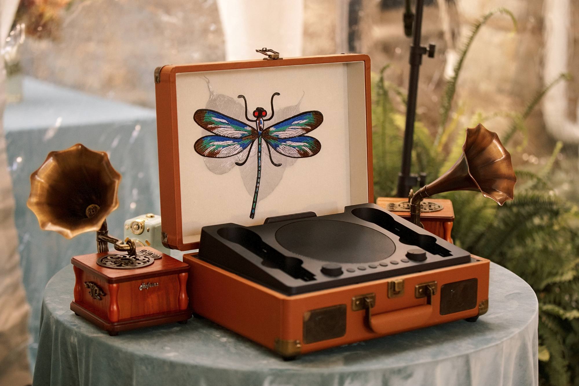 Vintage suitcase record player with an embroidered dragonfly in its lid, flanked by two small gramophone speakers on a table draped in pale blue velvet
