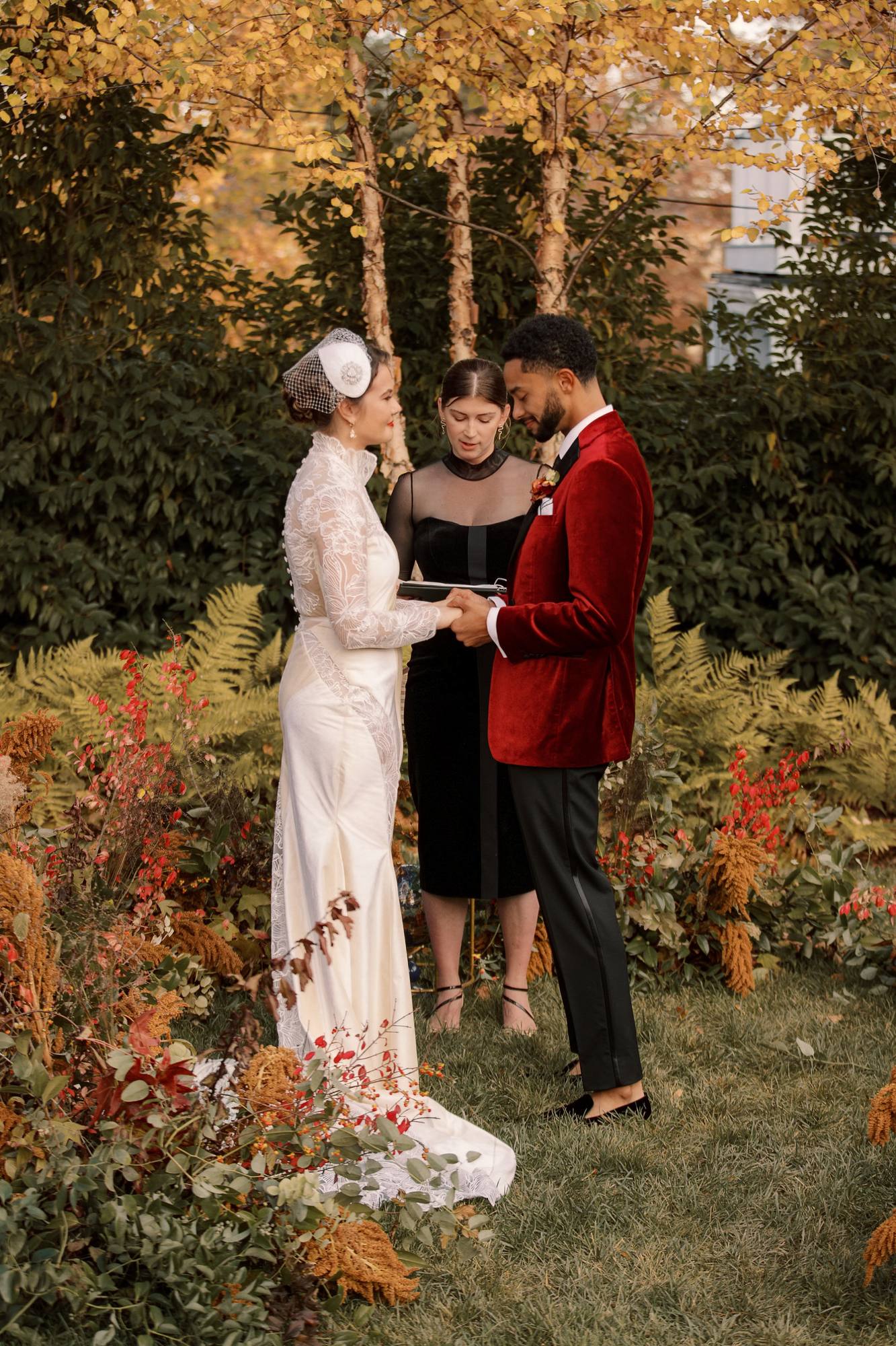 Bride and groom holding hands for their vows beneath yellow birch trees, with the officiant between them and low arrangements of red burning bush, golden amaranth and ferns