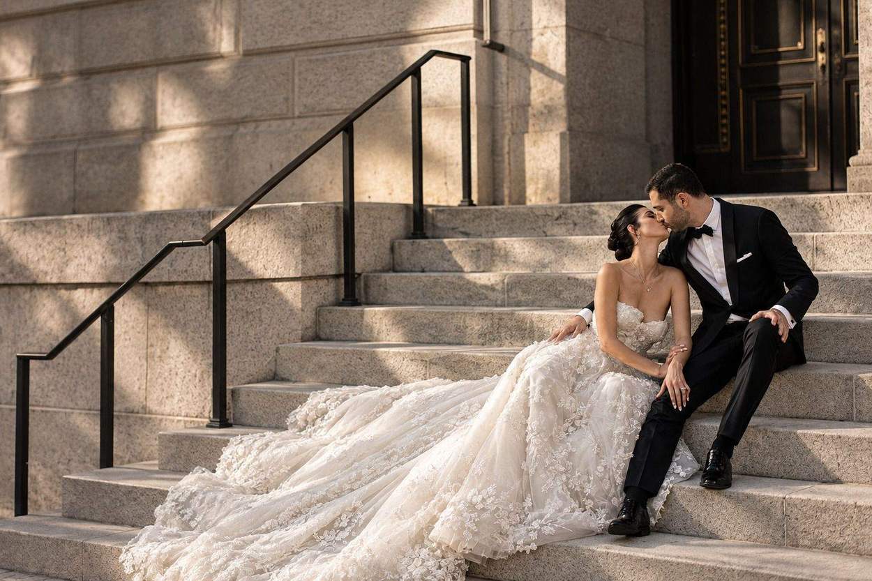 A bride in a floral-lace gown with a long train and a groom in a tuxedo kiss while seated on granite steps beside a black iron railing.