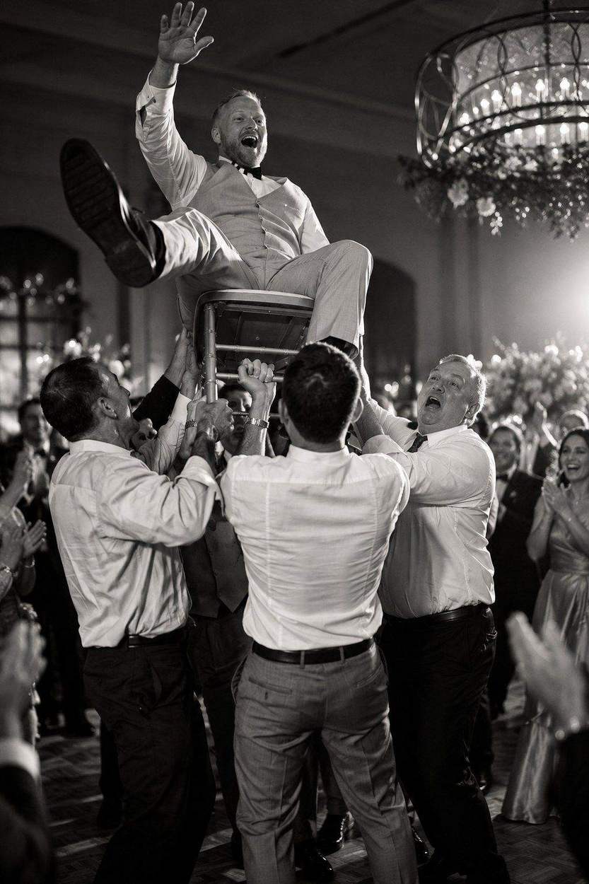 Washington DC photographer black and white wedding reception photo: a groom lifted high on a chair by cheering guests under a chandelier.